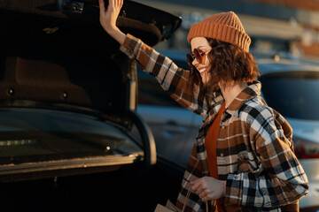 Woman opens car trunk with a smile in sunlight, beanie and sunglasses, casual shopping moment. Authenticity, candid lifestyle and golden hour glow evoke mindful living and emotional storytelling.