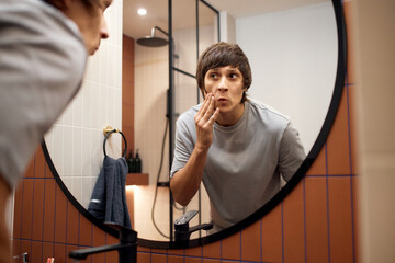 Portrait of young Caucasian man examining face in bathroom mirror, touching cheek with hand, appearing concerned while checking skin condition, modern shower visible in background