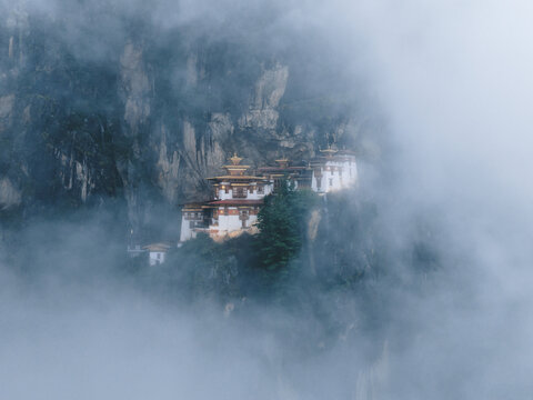 Aerial view of the iconic Tiger's Nest Monastery perched dramatically on a cliffside, shrouded in ethereal mists and framed by rugged terrain, Paro, Bhutan.