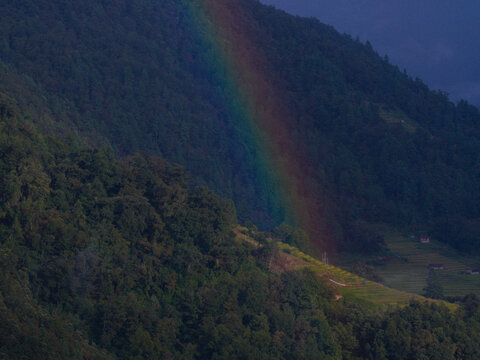 Aerial view of a vibrant rainbow arches over the lush, deep green valley, contrasting with the dark slopes and hinting at hidden villages, Bhutan.