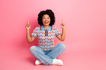 Happy stylish young woman sitting on pink studio background pointing up with fingers wearing a geometric patterned shirt and jeans
