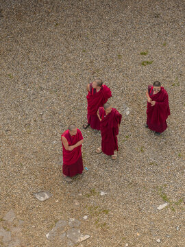 Aerial view of crimson-robed monks stand in contemplative silence on the gravel courtyard, their figures a striking contrast against the muted tones, Paro, Bhutan.