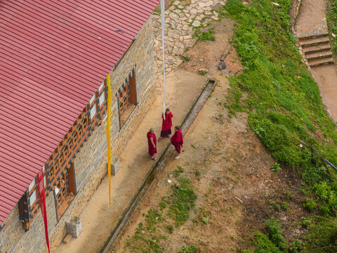 Aerial view of monks in deep red robes walking near a stone building with a vibrant red roof, amidst the lush greenery, Paro, Bhutan.
