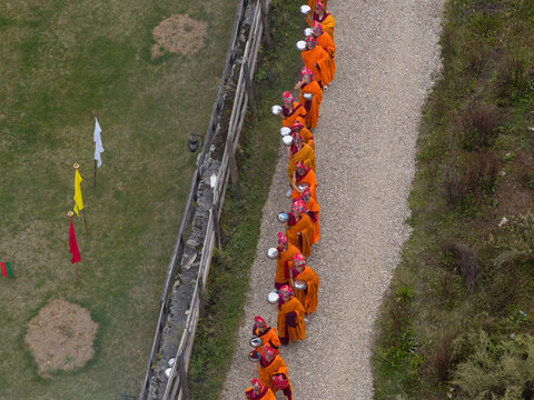 Gantey Monastery, Bhutan - 21 September 2025: Aerial view of monks in vibrant orange robes walking along a gravel path, a stark contrast to the bordering green grass and weathered stone wall.