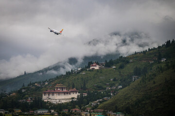 Paro, Bhutan - 18 September 2025: View of a vibrant aircraft soaring above the lush, green valley, juxtaposed against the backdrop of ancient temples and mist-shrouded mountains.