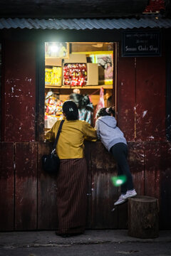 Haa, Bhutan - 25 September 2025: View of a woman and a girl peering into a brightly lit shop window near Lhakhang Karpo monastery, their forms contrasting with the shop's glow.