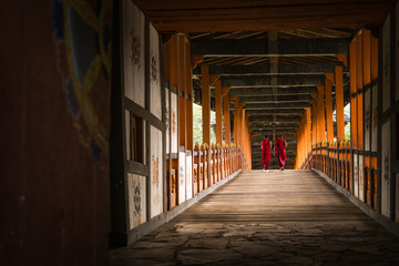 Punakha, Bhutan - 19 September 2025: View of two monks in vibrant red robes traverse the wooden bridge, framed by intricate patterns and warm hues of the structure.