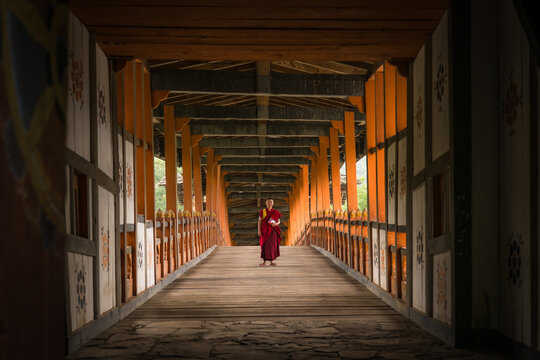 Punakha, Bhutan - 19 September 2025: View of a solitary monk in vibrant robes crossing the weathered wooden bridge, a passage framed by ornate carvings and the soft light of the valley.