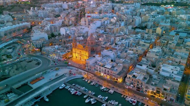 Marsaxlokk Malta fishing village harbor and illuminated parish church at twilight aerial drone view.
