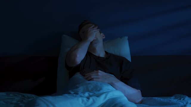 Tired young man lies awake in a dark bedroom, doom scrolling social media or news feed on his smartphone late at night and covering his face in frustration and exhaustion. Insomnia. Space for text 