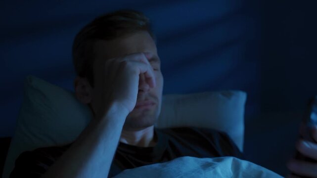 Young tired man with a phone in his hands scrolls through news feed or social media at night in his bedroom, but is interrupted by bad news or fatigue, disappointed and exhausted. 