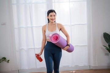 Woman carries yoga mat and water bottle in a bright room before starting a workout session