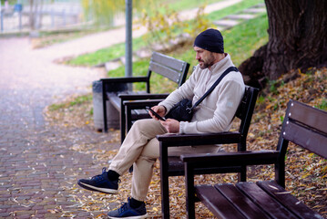 Fototapeta premium Man in a fleece jacket and beanie sitting on a park bench, absorbed in his smartphone. Autumn leaves cover the ground, creating a calm, reflective outdoor scene.