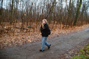 Obraz premium A thoughtful young woman walks alone on a quiet dirt path through a bare, autumnal forest, surrounded by fallen brown leaves and leafless trees under an overcast sky, carrying a book and backpack.