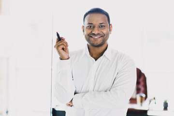 Young handsome businessman in a modern office smiling and presenting with a marker beside a glass board ready to share ideas and strategy