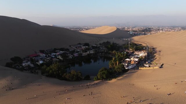 High drone descends to establish Huacachina oasis nestled in vast dunes with desert ridge lines in warm light