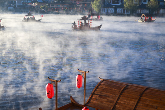 Serene, tranquil morning in traditional Chinese water town. Misty fog rises from river as people enjoy peaceful ride on an ancient wooden boat with red lanterns