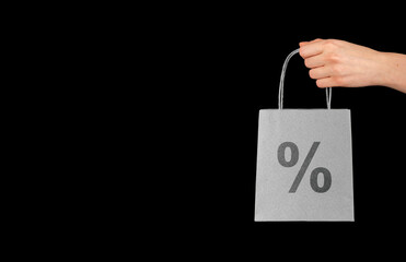 A hand holds a black shopping bag marked with a percentage sign, symbolizing a sale or discount. The