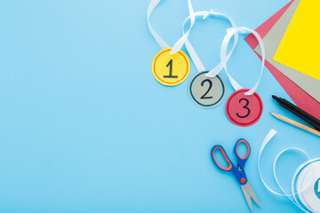 Created gold, silver and bronze paper medals with white ribbons. Scissors, marker, pencil and application paper on blue table background. Little child handcraft. Empty place for text. Top down view.