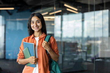 Portrait of a young smiling Indian female student standing indoors, holding a backpack and books,...