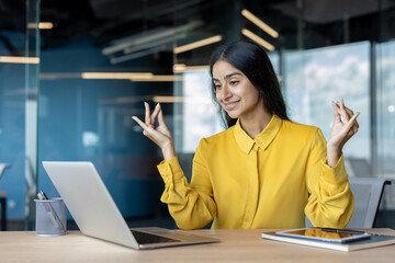 Smiling young Muslim businesswoman sitting at her desk in the office in a lotus position, looking at the laptop screen and meditating