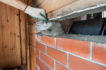 Bricklayer lays red brick around a sauna stove using a trowel. This is renovation work. The bricks are laid on a refractory sand-cement mixture.