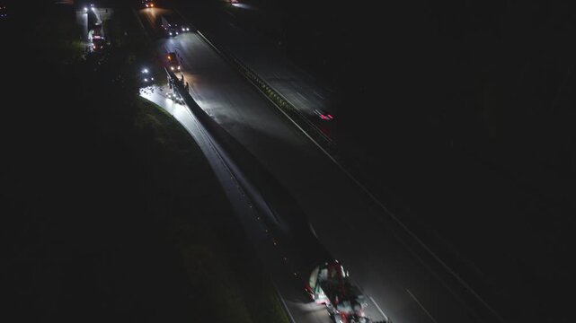Spectacular heavy transport of a wind turbine rotor blade at night on a multi-lane highway. The oversized truck is illuminated by escort vehicles on the dark road.
