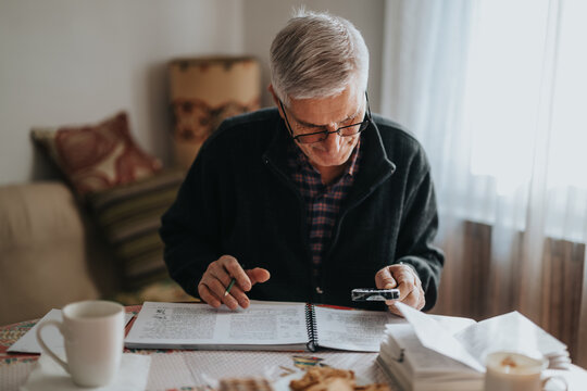 An elderly man sits at a dining table in a cozy home, focused on a spiral notebook and his smartphone. Surrounded by papers, a coffee mug, and light from a window, he concentrates on his work. - Powered by Adobe