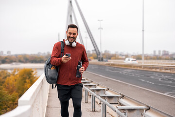 Man Walking on Bridge With Backpack and Smartphone, Smiling in Casual Attire