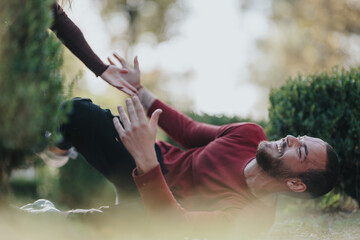 A bearded man in a red sweater lies on the grass in a sunny park, smiling as he reaches up for a high five with a friend. Casual, candid outdoor moment.