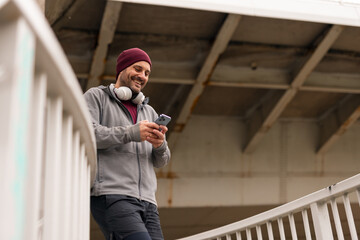 Man Smiling While Using Smartphone Outdoors Wearing Headphones and Beanie © Jelena