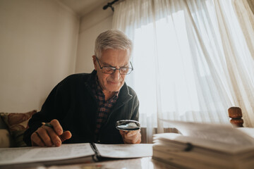 An elderly man with glasses leans over a desk, examining documents with a magnifying glass. He focuses intently in a sunlit home office, capturing a moment of study and quiet concentration.