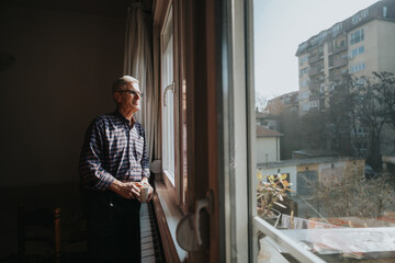 Older man stands by a sunlit window, holding a mug and gazing outside at an urban neighborhood. Warm indoor scene contrasts with the bright exterior, conveying calm, reflection, and everyday comfort.