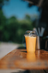 A refreshing orange smoothie served in a plastic cup, on a wooden table outdoors. The scene features a blurred natural background, creating a cozy and relaxed atmosphere on a sunny day.