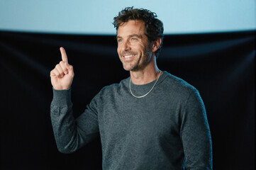 Fototapeta premium Man in gray sweater pointing upward with index finger while smiling against dark background