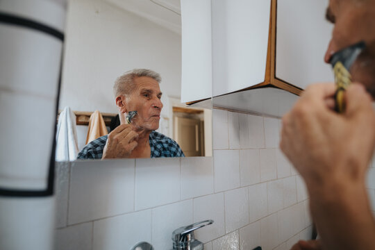 An elderly man stands at a bathroom sink, applying shaving cream and shaving with a razor, reflected in the mirror. A quiet, private moment of daily grooming and self-care. - Powered by Adobe