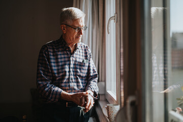 An elderly man in a plaid shirt stands by a sunlit window, hands resting as he gazes outside. A mug sits on the sill, conveying calm, reflection and everyday domestic life.