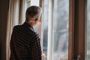 An older man stands at a sunlit window, gazing outside with a thoughtful expression. He wears a...