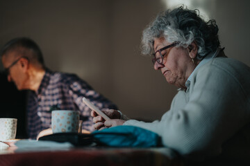 A senior woman sits at a table, focused on her smartphone, wearing a light shirt and glasses. A blurred man in the background adds a sense of family life and a quiet morning routine.