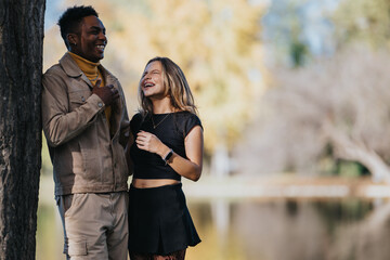 Friends share a joyful moment outdoors, leaning against a tree near a tranquil lake. The autumn colors, casual outfits, and warm smiles convey friendship, relaxation, and carefree moments in nature.