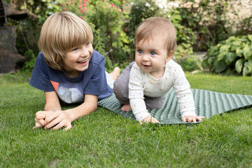 Two children, an older brother and a younger sister, lie on the grass in the garden, happily playing with each other, exploring the world around them. Summer walks, babysitting. Childhood moments