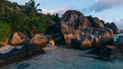 Couple walking on a tropical beach among large granite rocks at sunset and palm trees