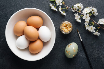 Top view flat lay of Easter egg decorating. Natural eggs, gold leaf, a paintbrush, and spring blossoms on a dark, textured background.