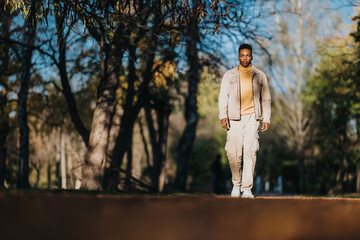 A stylish man walks along a sunlit park path, wearing a beige jacket and mustard turtleneck. Warm tones and natural light convey a relaxed, casual outdoor vibe and contemporary street style.