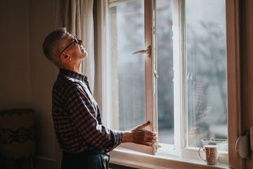 A senior man in a plaid shirt stands by a sunlit window, looking outside with a contemplative mood. A coffee mug rests on the sill, capturing a quiet, cozy home moment.