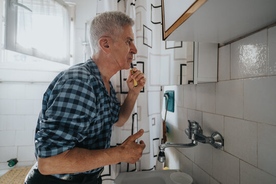 An older man in a checkered blue shirt brushes his teeth at a bathroom sink, illuminated by natural light. The scene captures daily hygiene, self-care, and a calm morning routine.