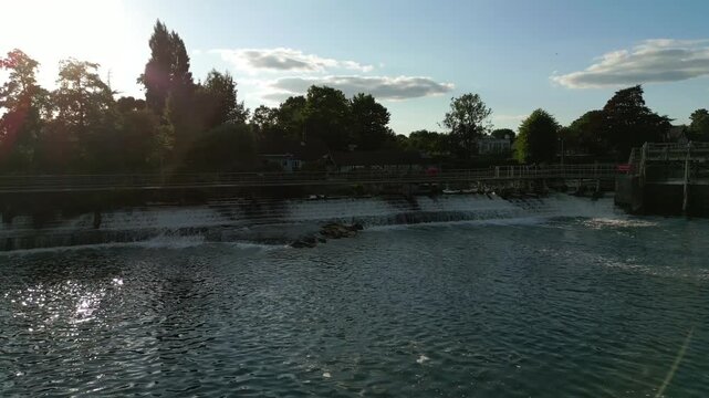 A serene river with a waterfall, bridge, and trees under a blue sky with clouds.