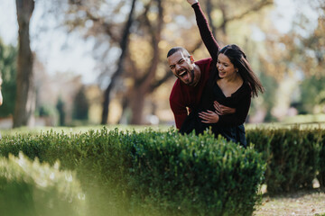 A happy couple in casual wear pose behind a hedge, laughing and embracing in a sunlit park. A candid moment of love, friendship, and carefree outdoor joy.