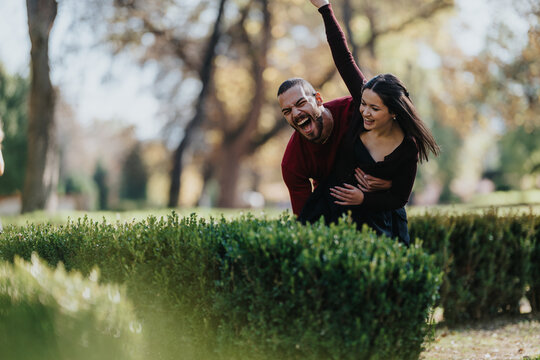 A happy couple in casual wear pose behind a hedge, laughing and embracing in a sunlit park. A candid moment of love, friendship, and carefree outdoor joy. - Powered by Adobe