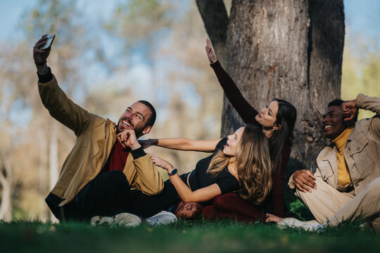 A diverse group of friends lounges on the grass by a large tree, capturing a fun selfie together. The sunny park setting conveys friendship, happiness, and relaxed outdoor moments. - Powered by Adobe